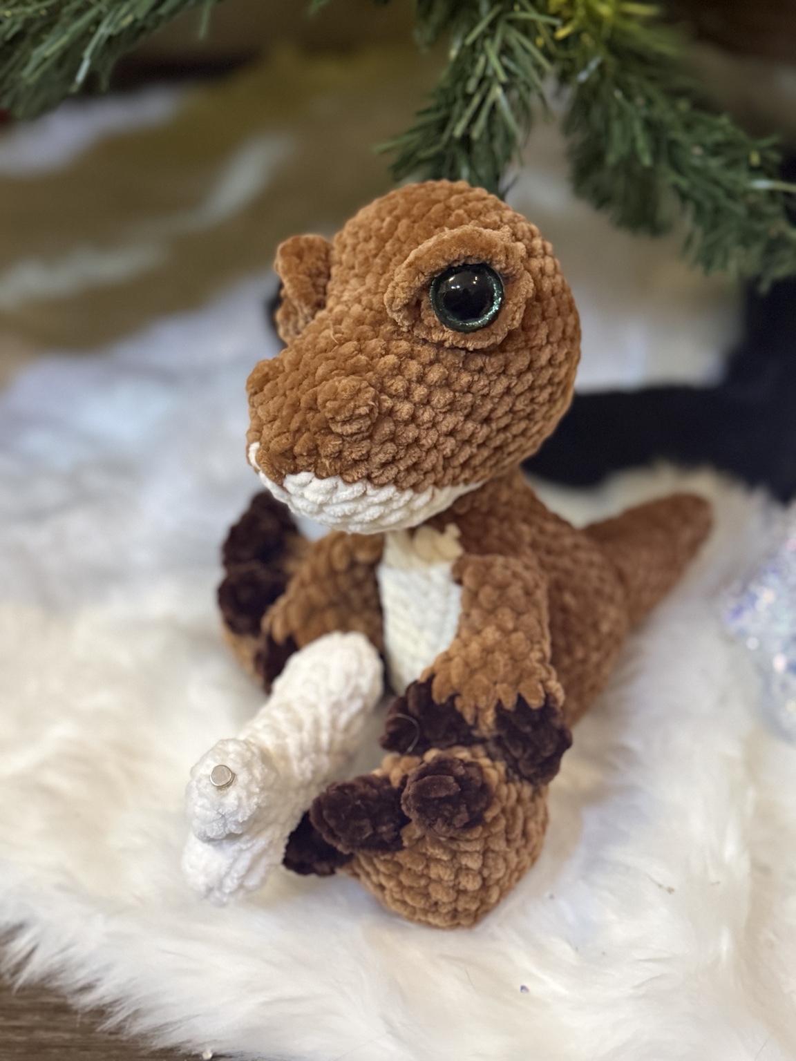 Brown crochet dinosaur with cream details beside a Christmas tree.