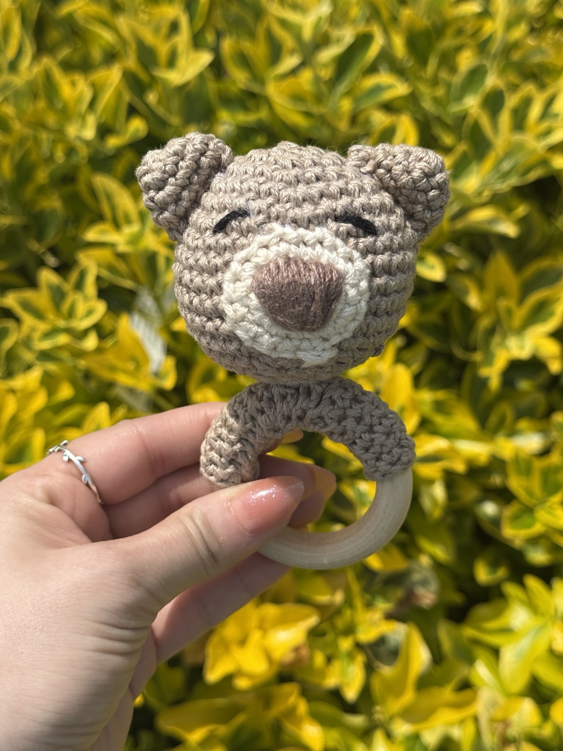 Small gray crochet bear with a wooden ring, held in front of green leaves.
