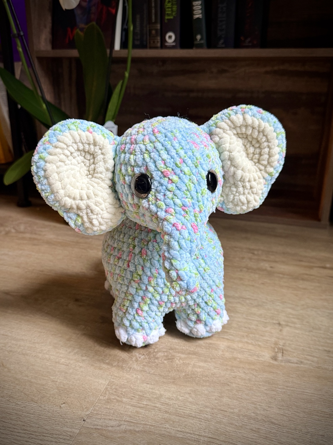Pastel multicolored crochet elephant with cream ears, standing on a wooden table.