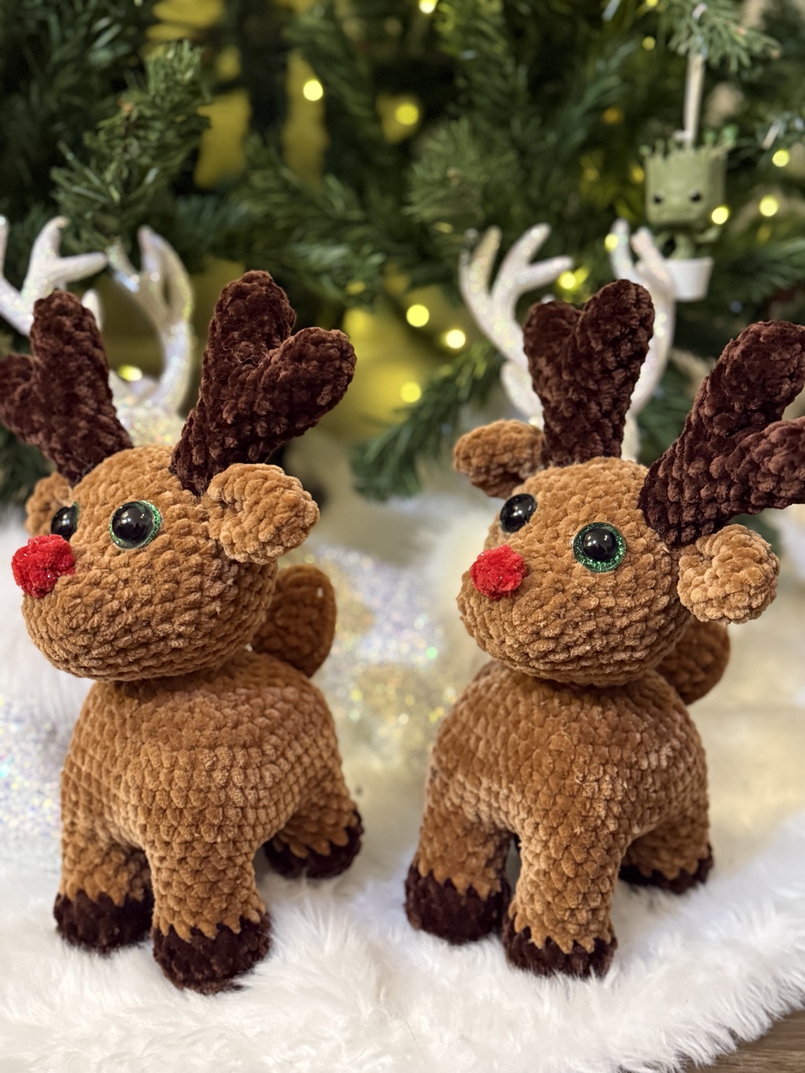 Two brown crochet reindeer with red noses and dark antlers, beside a Christmas tree.