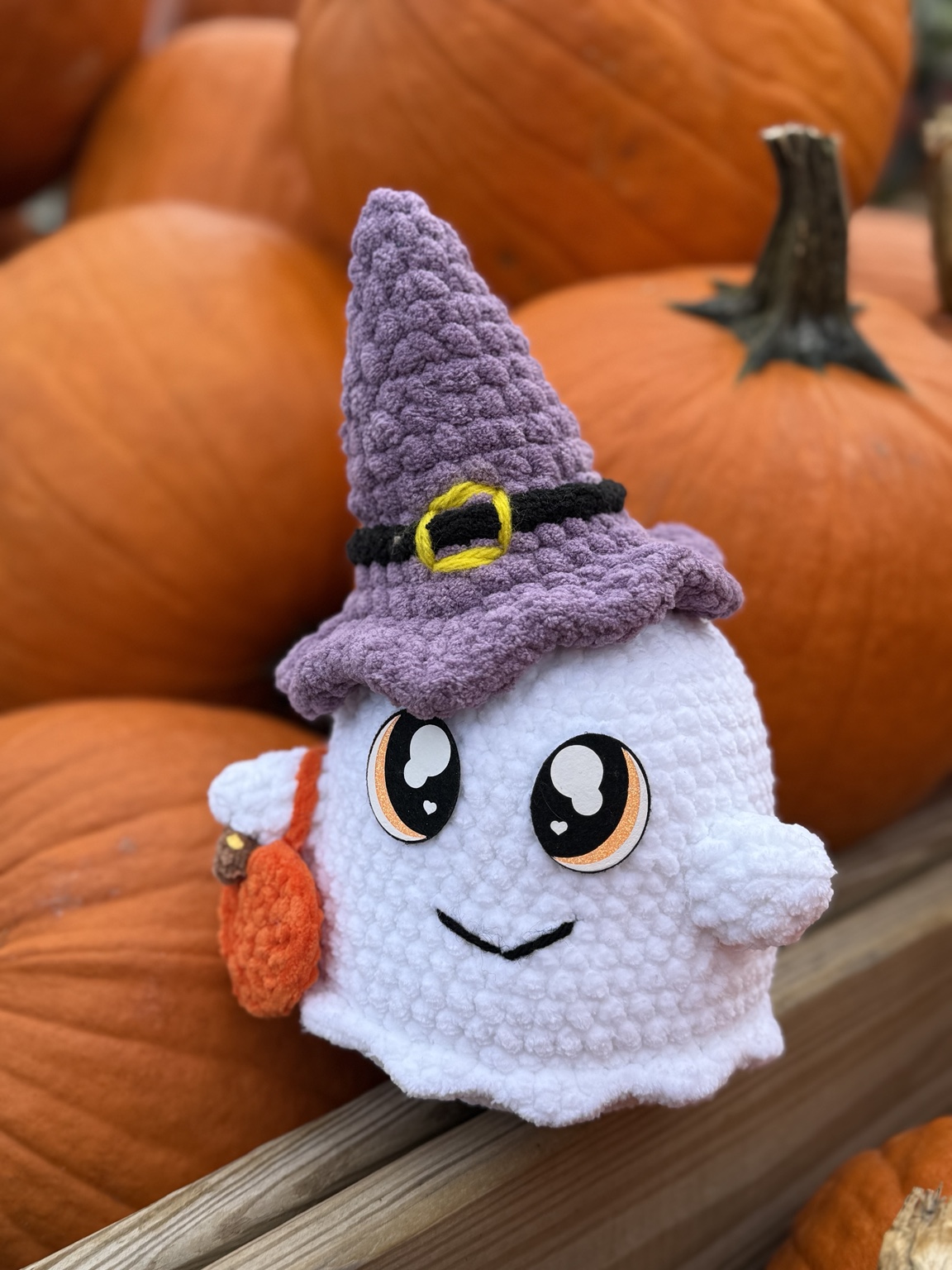 White crochet ghost with a purple witch hat and orange bag, sitting among pumpkins.